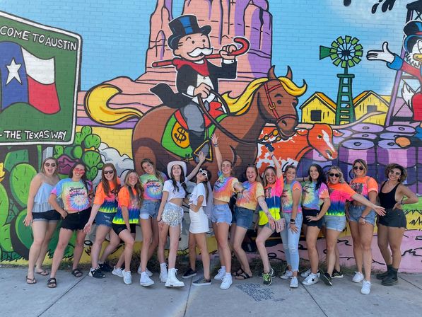 Group of women in colorful tie-dye shirts and summer outfits posing for a fun group photo in front of a vibrant Austin street mural featuring a Texas flag, cartoon top-hat rider on a horse, and desert farm imagery.