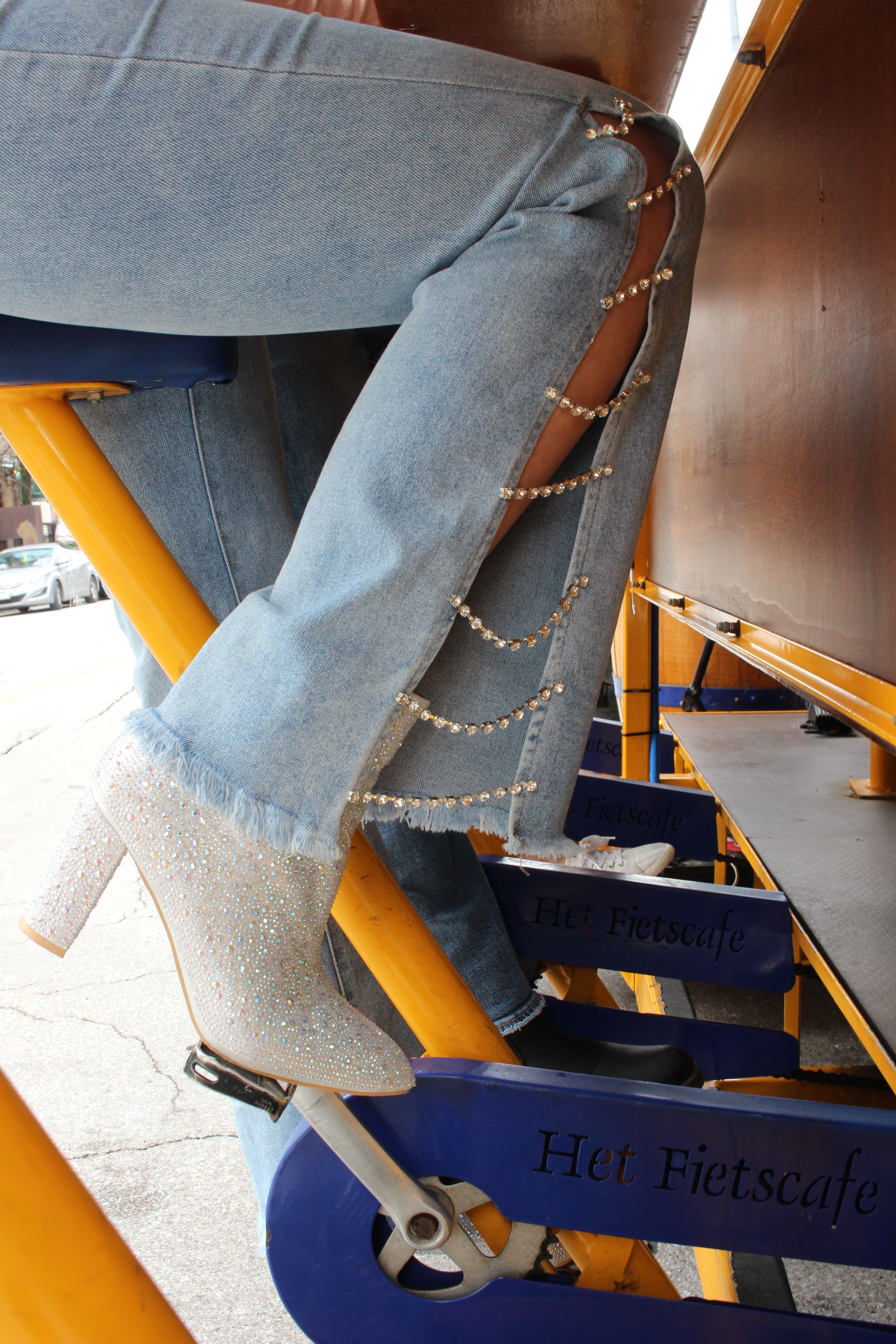 Sparkly rhinestone high-heeled ankle boot resting on a pedal of a yellow outdoor pedal-powered bar, light-wash jeans with side cutouts linked by crystal chains.