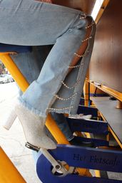 Sparkly rhinestone high-heeled ankle boot resting on a pedal of a yellow outdoor pedal-powered bar, light-wash jeans with side cutouts linked by crystal chains.