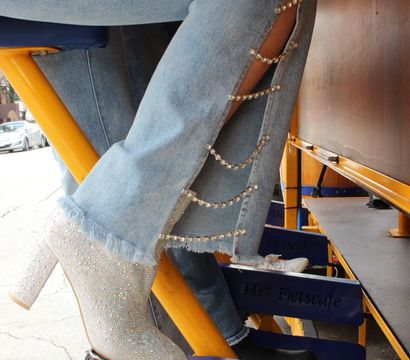 Sparkly rhinestone high-heeled ankle boot resting on a pedal of a yellow outdoor pedal-powered bar, light-wash jeans with side cutouts linked by crystal chains.