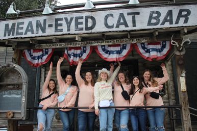 Group of seven women in matching peach shirts and jeans cheerfully wave on the porch of a rustic western bar storefront decorated with red-white-blue bunting, weathered wood, a cowboy hat, and mounted antlers.