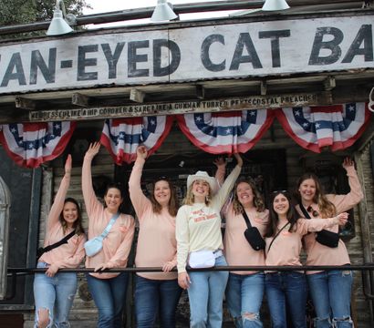 Group of seven women in matching peach shirts and jeans cheerfully wave on the porch of a rustic western bar storefront decorated with red-white-blue bunting, weathered wood, a cowboy hat, and mounted antlers.