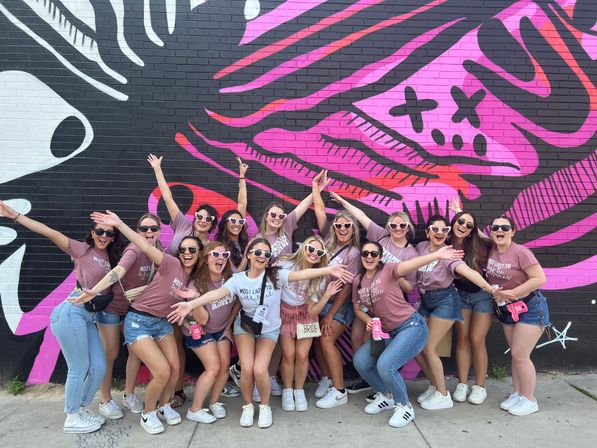 Bachelorette party group of women in matching mauve shirts and heart-shaped sunglasses, including the bride in a white tee and 'Bride' bag, posing joyfully in front of a vibrant pink and black street-art mural on a brick wall.