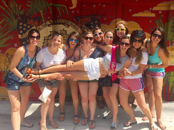 Smiling group of women at a bachelorette party holding the bride-to-be horizontally in front of a vibrant tropical mural on a sunny street, wearing sunglasses and summer outfits