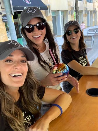 Three smiling women at an outdoor patio counter wearing matching gray baseball caps with a Texas outline and sunglasses, one holding a colorful iridescent disco-ball cup with straw.