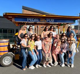 Smiling group of women posing by a pedal-powered party bike on a sunny urban street, wearing sunglasses and casual summer outfits.