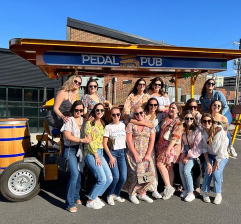 Smiling group of women posing by a pedal-powered party bike on a sunny urban street, wearing sunglasses and casual summer outfits.
