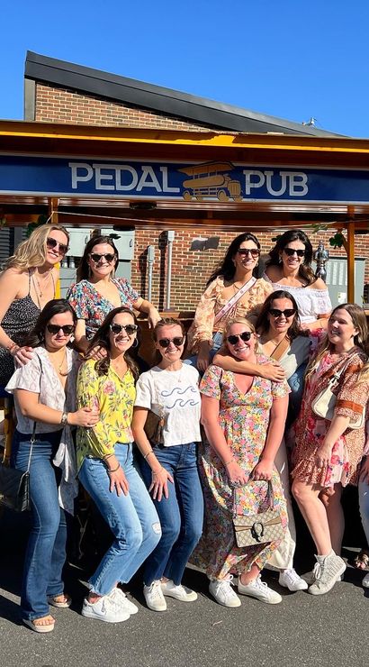 Smiling group of women posing by a pedal-powered party bike on a sunny urban street, wearing sunglasses and casual summer outfits.