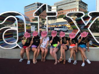 Seven friends, one wearing a 'Bride' shirt, sit on a bench in front of a large colorful 'ATX' sign in Austin, holding pink cowboy hats over their faces for a playful bachelorette-photo moment.