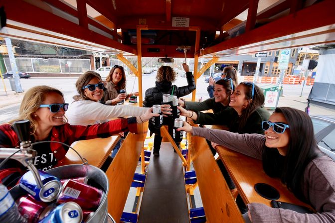 Group of friends wearing sunglasses toasting canned drinks aboard a bright orange pedal pub party bike on a downtown city street during daytime