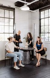 Sunlit industrial-style studio with large paned windows — two clinicians set up IV drips behind a round pedestal table while two clients sit nearby