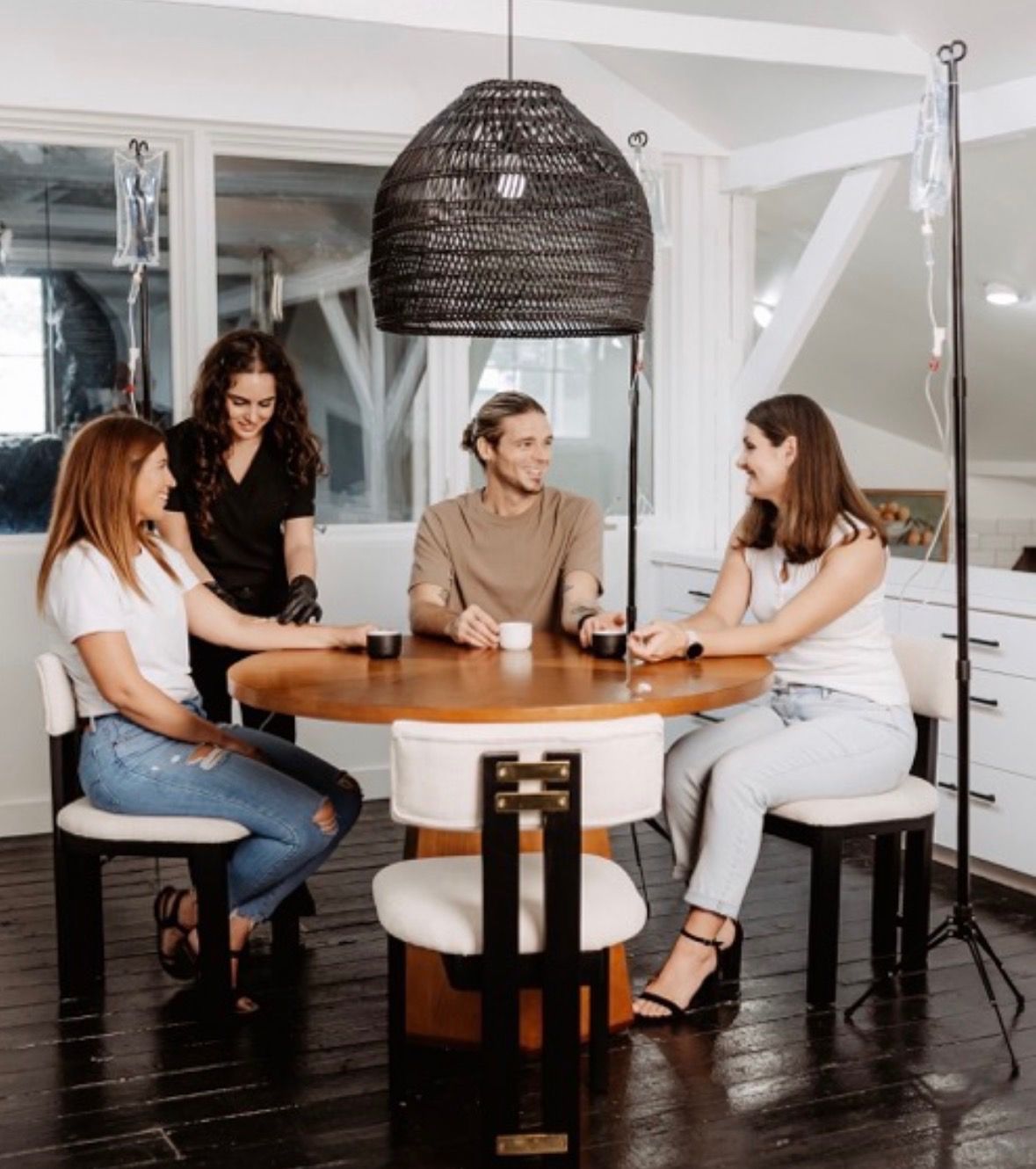 Four adults seated around a round wooden table in a bright loft-style wellness studio, chatting and sipping while two IV bags on poles hang nearby under a woven pendant light.