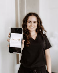 Smiling medical professional in black scrubs holds a smartphone toward the camera showing a mobile wellness app logo in a clinic setting