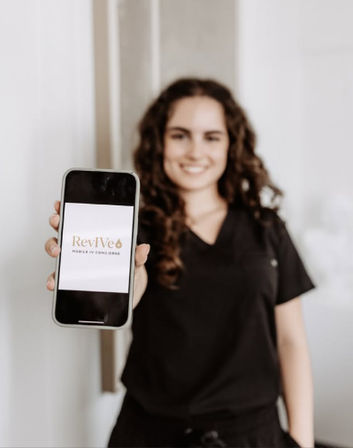 Smiling medical professional in black scrubs holds a smartphone toward the camera showing a mobile wellness app logo in a clinic setting