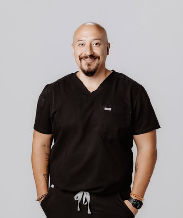 Smiling bald medical professional in black scrubs with a goatee, visible arm tattoos and smartwatch, hands in pockets against a neutral gray studio background — friendly clinical portrait.