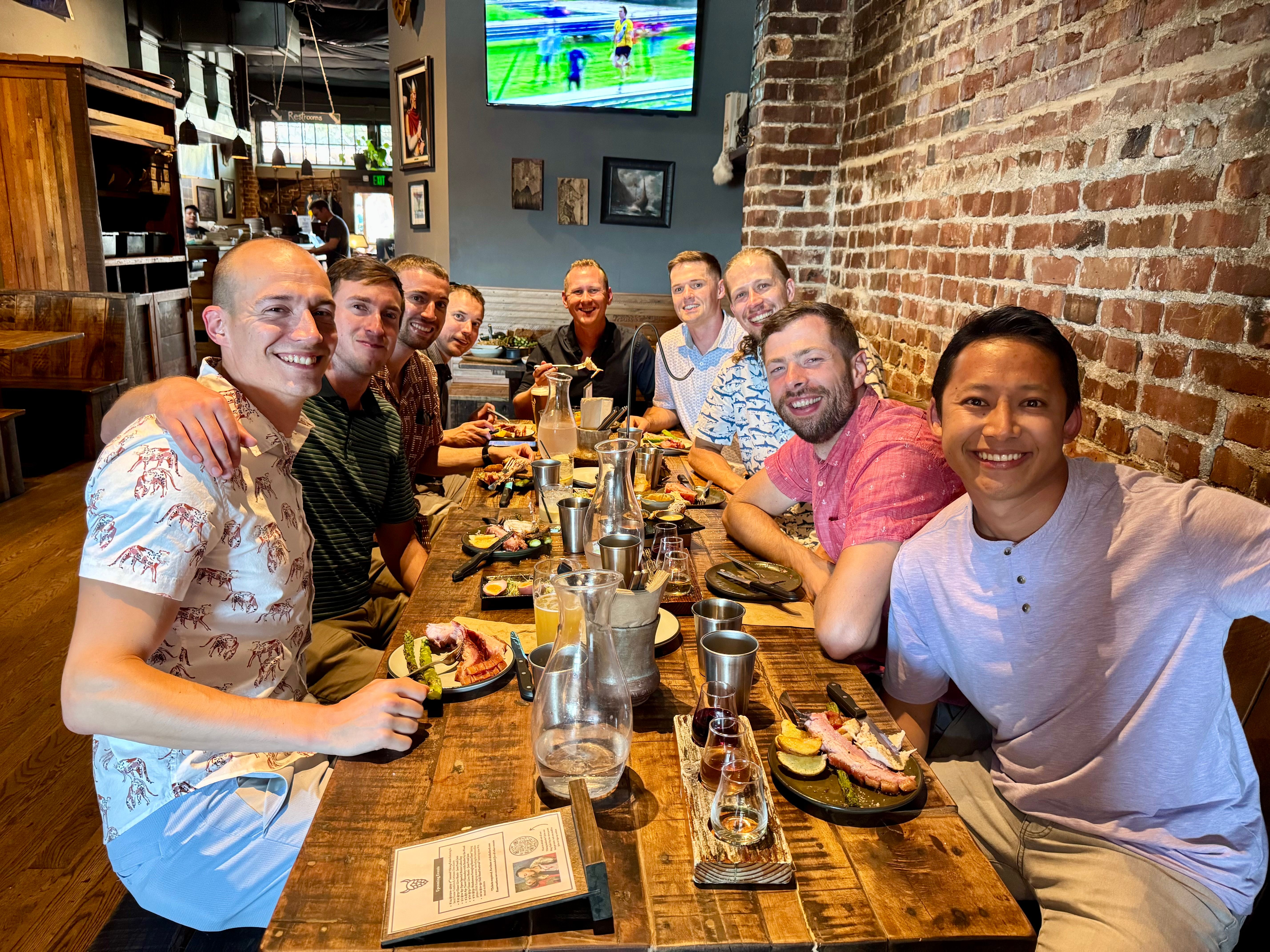 Nine friends smiling at a long communal wooden table in a rustic brick‑walled restaurant, sharing plates, drinks, and laughter during a casual group dinner.