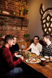 Four friends enjoying a cozy meal at a brick-walled bistro, sharing a rustic wooden platter of grilled fish, skewers and sides at a warmly lit table with potted plants and craft mugs.