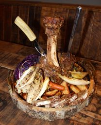Rustic meat platter with a tall roasted bone-in beef shank standing upright on a wooden board, surrounded by grilled endive, purple cabbage, roasted carrots, potato wedges and crusty bread