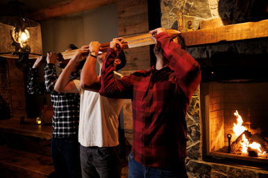 Three friends in flannel taking a group shot from a wooden shot-ski beside a roaring stone fireplace in a cozy rustic lodge.