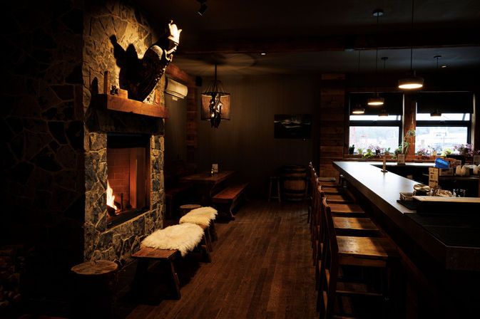 Cozy rustic bar interior with a lit stone fireplace, sheepskin-covered bench, long wooden bar lined with stools, warm pendant lighting and a window view.