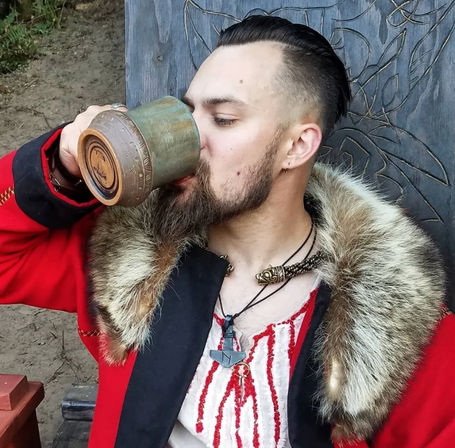 Person in a fur-trimmed red coat and embroidered tunic sipping from a rustic ceramic tankard, wearing braided cord and pendant necklaces — outdoor historical reenactment vibe.