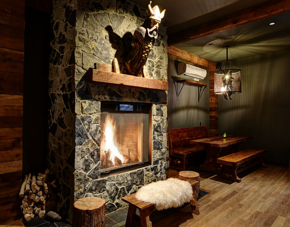 Cozy rustic mountain-lodge interior featuring a stone fireplace with roaring fire, carved wooden bust on the mantel, wooden bench and dining table, log stools and sheepskin seat, warm pendant lighting and stacked firewood.