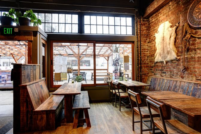 Cozy rustic cafe interior with reclaimed wood tables and benches, exposed brick wall with a pale animal hide, large industrial windows overlooking a covered outdoor patio with pergola seating.