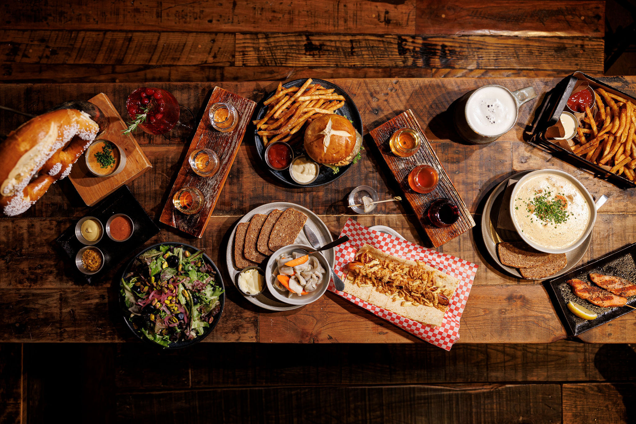 Overhead shot of a rustic wooden table with a mouthwatering pub-style spread: burger and fries, soft pretzel with dip, mixed green salad, creamy chowder with rye, long sandwich on red-check paper, grilled fish, dark bread with pickled sides, a beer flight and frothy beer mug