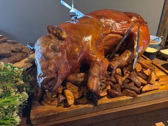 Whole roasted pig with shiny brown skin on a wooden carving board, surrounded by roasted potato wedges, tongs and nearby green vegetable side and dipping sauces