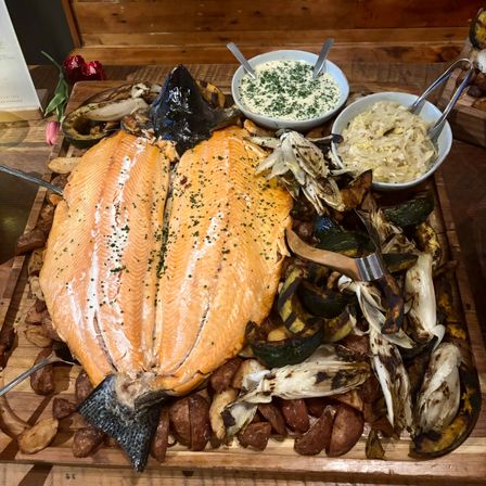 Whole roasted salmon on a wooden serving board with roasted potatoes, charred squash and endive, creamy herb dip and braised cabbage, a shareable seafood platter.