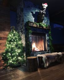 Cozy rustic cabin living room with a lit Christmas tree, stone fireplace with roaring fire and evergreen garland, carved figure wearing a Santa hat, fur-covered bench and log stool — warm holiday decor.