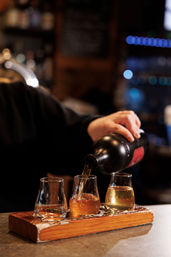 Bartender pouring whiskey into a three-glass tasting flight on a rustic wooden paddle at a cozy bar with warm bokeh lights