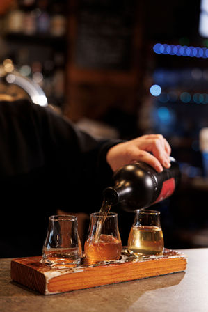 Bartender pouring whiskey into a three-glass tasting flight on a rustic wooden paddle at a cozy bar with warm bokeh lights