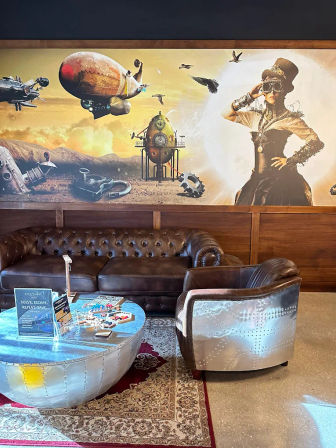 Steampunk-themed lounge with tufted brown leather sofa, riveted metal armchair and round riveted metal coffee table beneath a large airship and aviator mural.