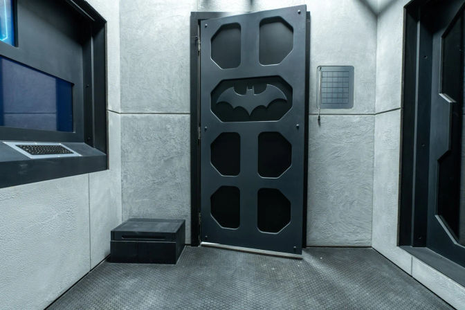 Batman-themed escape room interior with a matte black metal door cut with a bat emblem, textured gray concrete walls, wall-mounted grid panel, small black step box and a keypad window on rubberized flooring