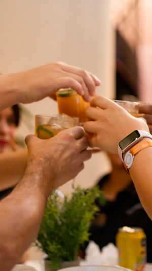 Close-up of friends toasting iced citrus cocktails with lime garnish at a cozy indoor restaurant — hands, bracelets and a smartwatch visible.