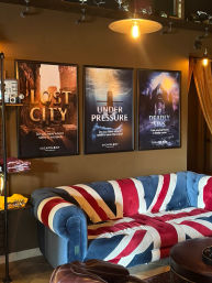 Union Jack upholstered sofa in a cozy escape-room lounge with three framed adventure posters titled 'Lost City', 'Under Pressure', and 'Deadly Sins' under warm industrial pendant lighting.