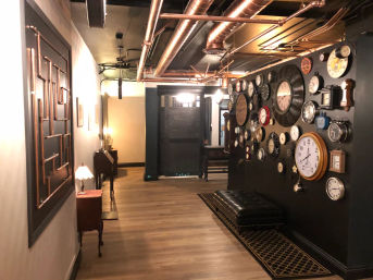 Interior hallway with exposed copper pipes and hardwood floor, black accent wall covered in assorted vintage clocks and a tufted leather bench — steampunk-inspired hotel corridor.