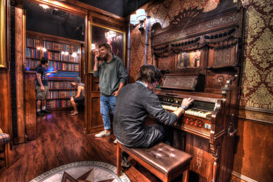 Cozy wood-paneled parlor with an ornate carved antique pump organ being played, compass motif on the hardwood floor, and a small library nook through a doorway where visitors browse books under warm sconces.