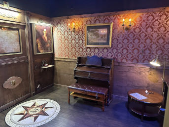Victorian-style parlor interior with antique upright piano and tufted bench, red damask wallpaper, framed portraits and brass wall sconces, compass medallion on hardwood floor, round side table with lamp and pipe.