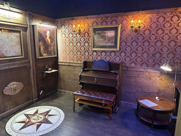 Victorian-style parlor interior with antique upright piano and tufted bench, red damask wallpaper, framed portraits and brass wall sconces, compass medallion on hardwood floor, round side table with lamp and pipe.