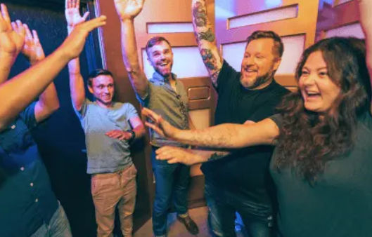Five adults laughing and raising their hands in a colorful indoor game room, celebrating a shared win.