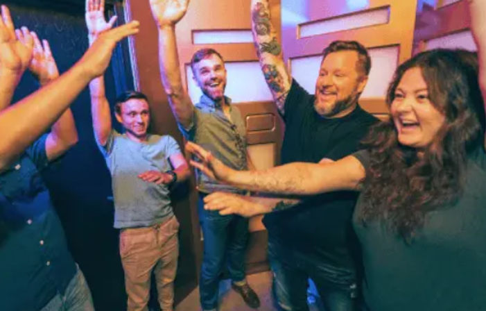 Five adults laughing and raising their hands in a colorful indoor game room, celebrating a shared win.
