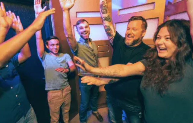 Five adults laughing and raising their hands in a colorful indoor game room, celebrating a shared win.