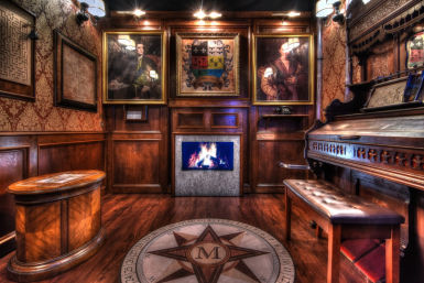 Warm wood‑paneled Victorian-style parlor interior with a glowing fireplace, framed portraits and heraldic crest, ornate pump organ and leather bench, and a compass-rose floor medallion.
