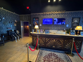 Dimly lit themed escape room lobby reception with carved wooden front desk, patterned wallpaper, wall-mounted screens, lamps, red velvet stanchions, a skeleton prop seated in a chair, posters and a decorative rug.