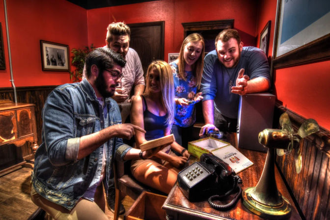 Five friends gathered around a wooden desk in an escape room, excitedly examining a clue box and book next to a vintage telephone and brass fan.