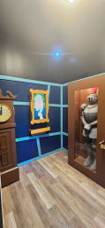 Playful fantasy-themed indoor exhibit room with blue paneled walls, a wooden grandfather clock, a framed cartoon portrait, a glass-encased suit of knight armor and light wood plank flooring.