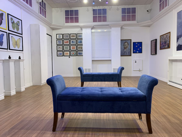 Bright white art gallery interior exhibition space with wood floors, framed artworks on the walls and two blue tufted benches facing each other, butterfly prints visible on the left wall.