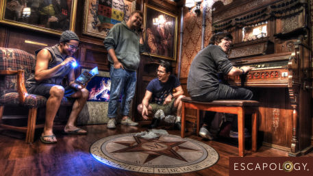 Four adults solving an ornate Victorian-style escape room study: one reads a clue by flashlight, one on a phone, one crouches by a compass floor medallion, another plays an antique organ by a faux fireplace.